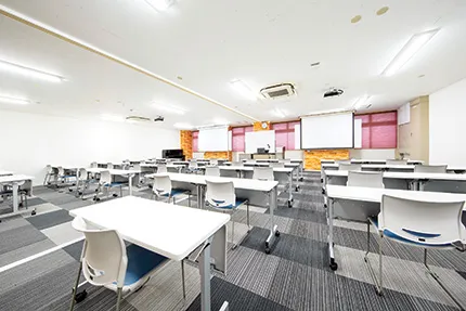 こども學舎の第1.2教室。広い空間で前にはホワイトボードやスクリーンがあり、たくさんの机が並ぶ。